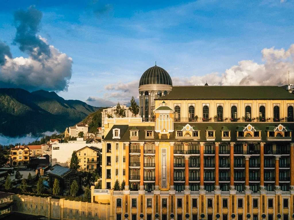 A grand hotel with a green dome, yellow and white facade, and multiple windows, set against a mountain landscape with a blue sky and clouds.
