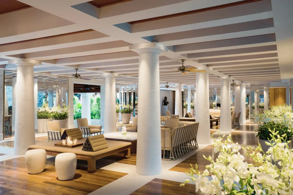 Spacious indoor lounge area with white pillars, ceiling fans, and wooden flooring. Comfortable chairs, sofas, tables, and abundant white flowers create a relaxing atmosphere with natural light coming through large windows.