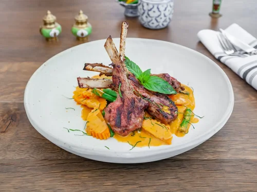 Grilled lamb chops served on a white plate with mashed sweet potatoes and green beans, garnished with fresh basil and herbs.
