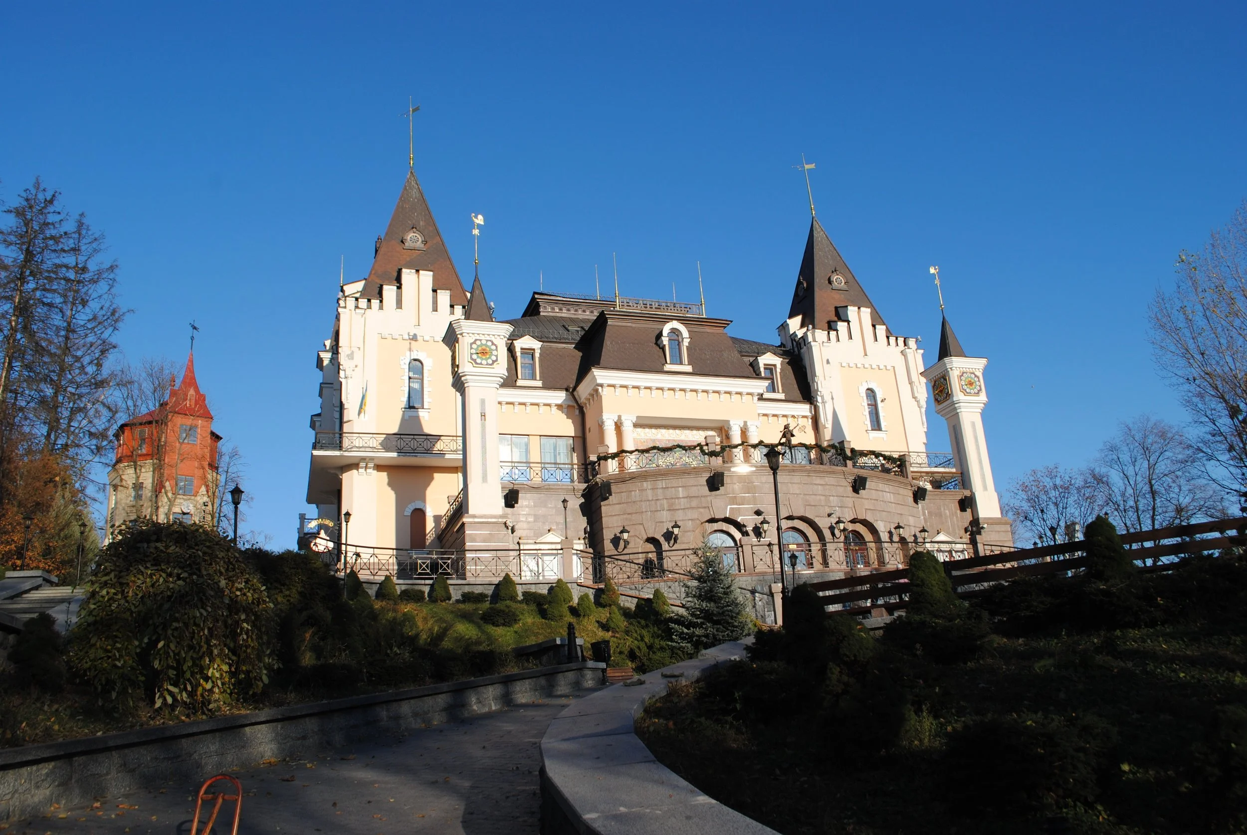 A castle-like building with multiple towers and flags, surrounded by trees and a landscaped garden under a clear blue sky.