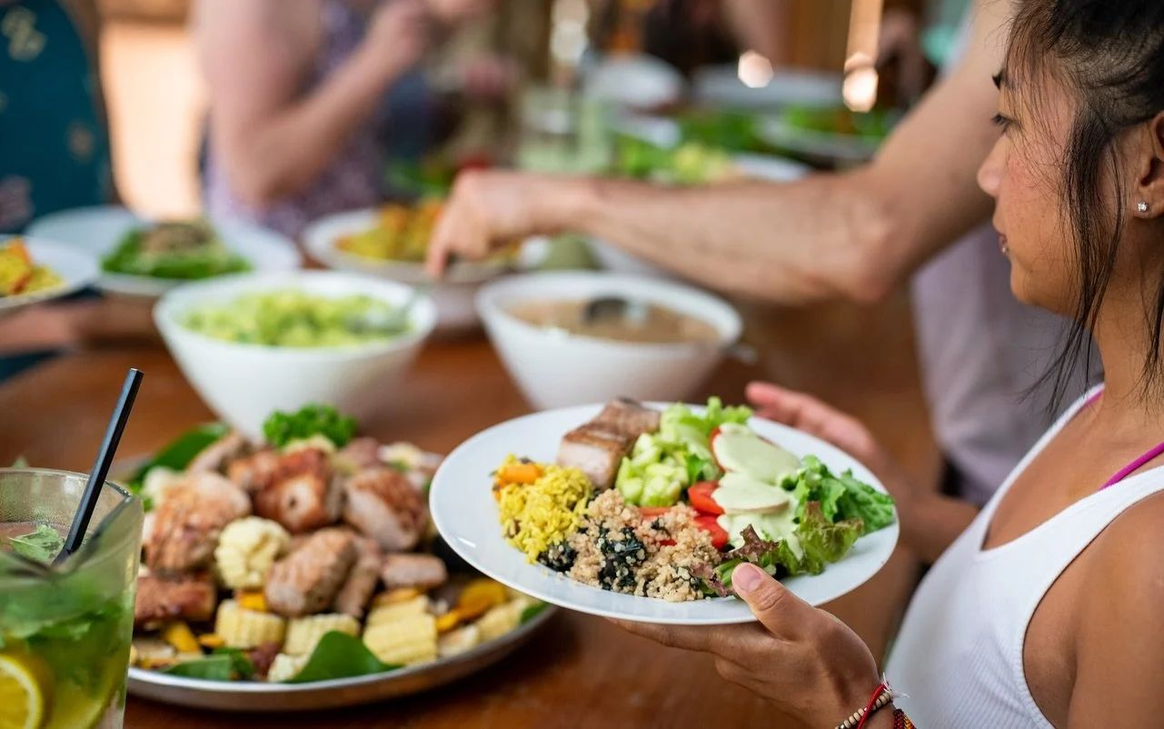 Woman holding a plate of salad with vegetables, grains, and a piece of meat at a table with a variety of food dishes.