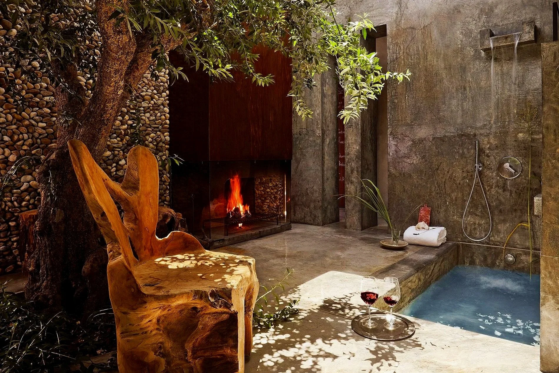 Indoor relaxation area with a tree growing next to a stone fireplace with a fire burning. There is a rustic wood chair in the foreground, a towel on a tray, and two glasses of red wine near a pool or hot tub. The area features stone and concrete walls and modern fixtures.