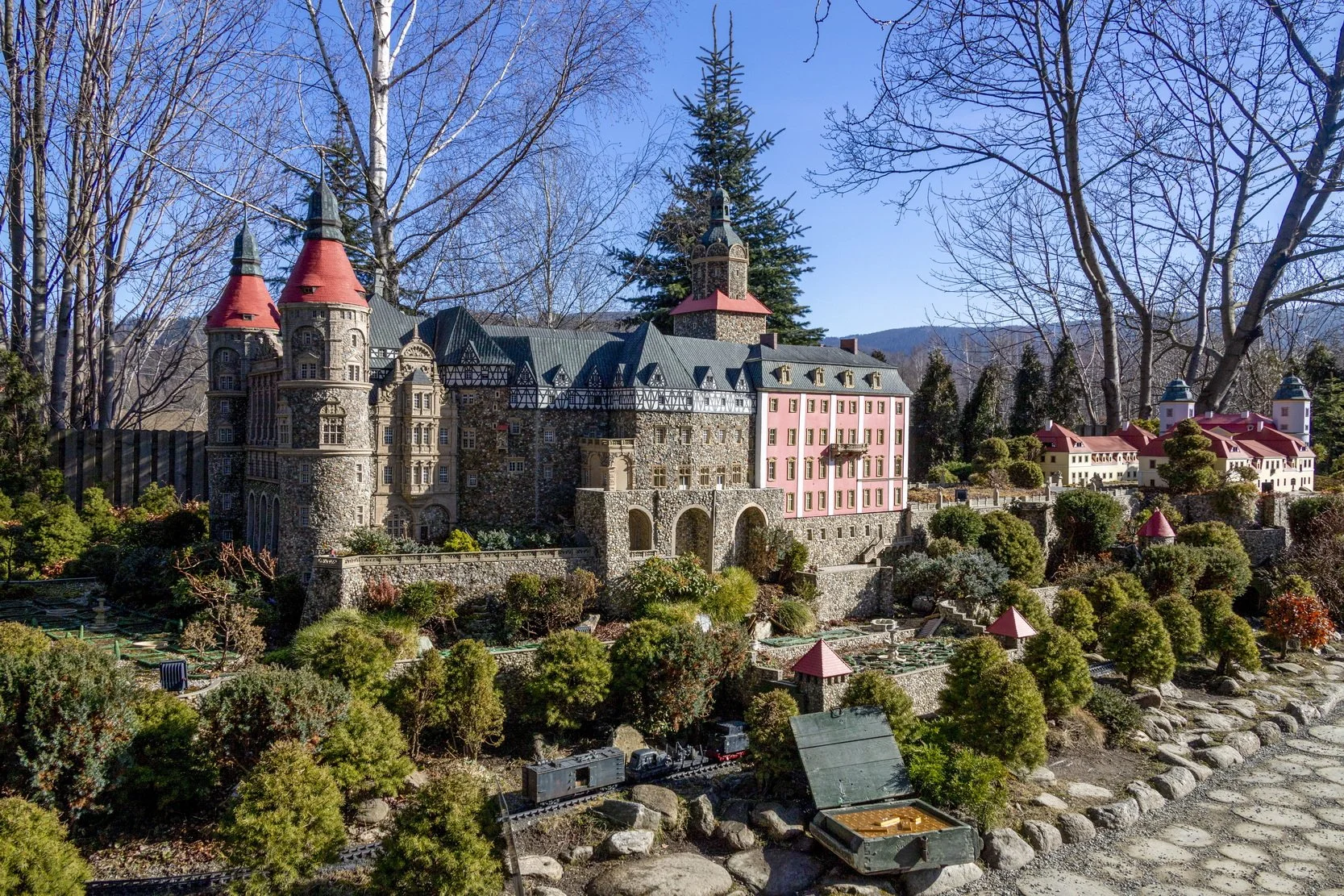 Model of a fairy tale castle with towers, turrets, a pink section, and a landscaped garden with trees and plants, set in a mountainous outdoor area.