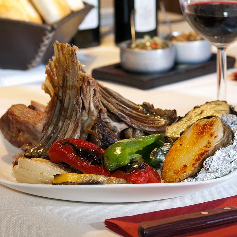 A plate of cooked lamb ribs with grilled vegetables, including red and green bell peppers, and roasted potato slices, on a table set with a glass of red wine.
