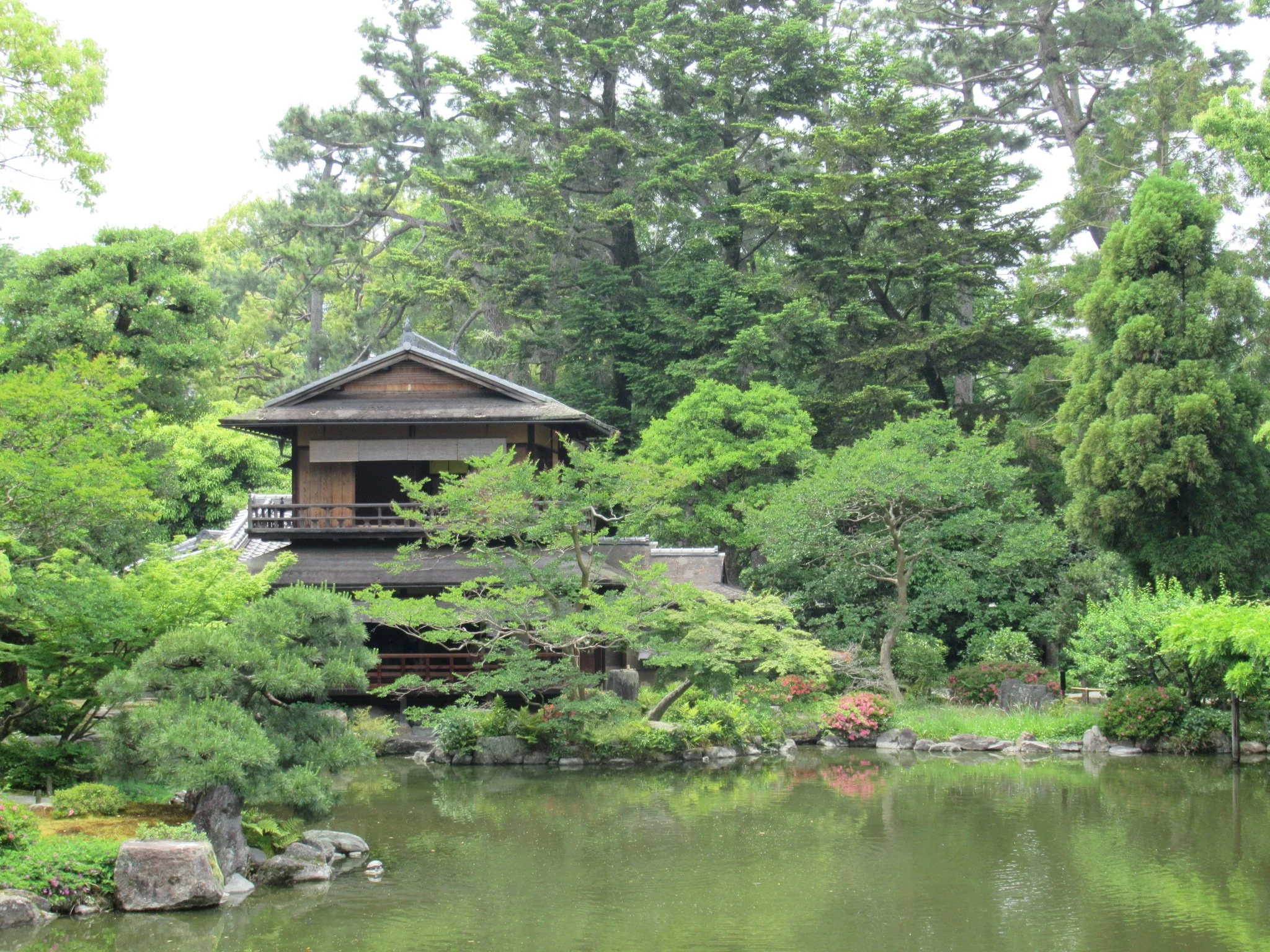 A traditional Japanese wooden pavilion with a tiled roof, surrounded by lush green trees and a pond in a Japanese garden.