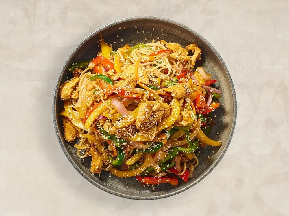 A bowl of stir-fried noodles with vegetables and chicken, topped with sesame seeds.