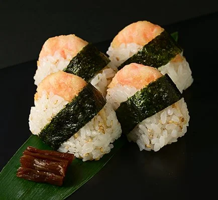 Four pieces of shrimp nigiri sushi with seaweed strip, served on a green leaf with a small portion of brown sauce.
