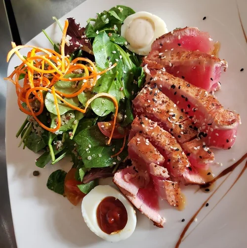 Mixed green salad with shredded carrots, cherry tomato, sliced seared tuna topped with sesame seeds, and a halved soft-boiled egg on a white plate.