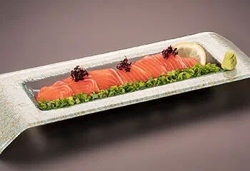 Sliced salmon sashimi on a rectangular dish garnished with green onions, black caviar, lemon wedge, and wasabi.