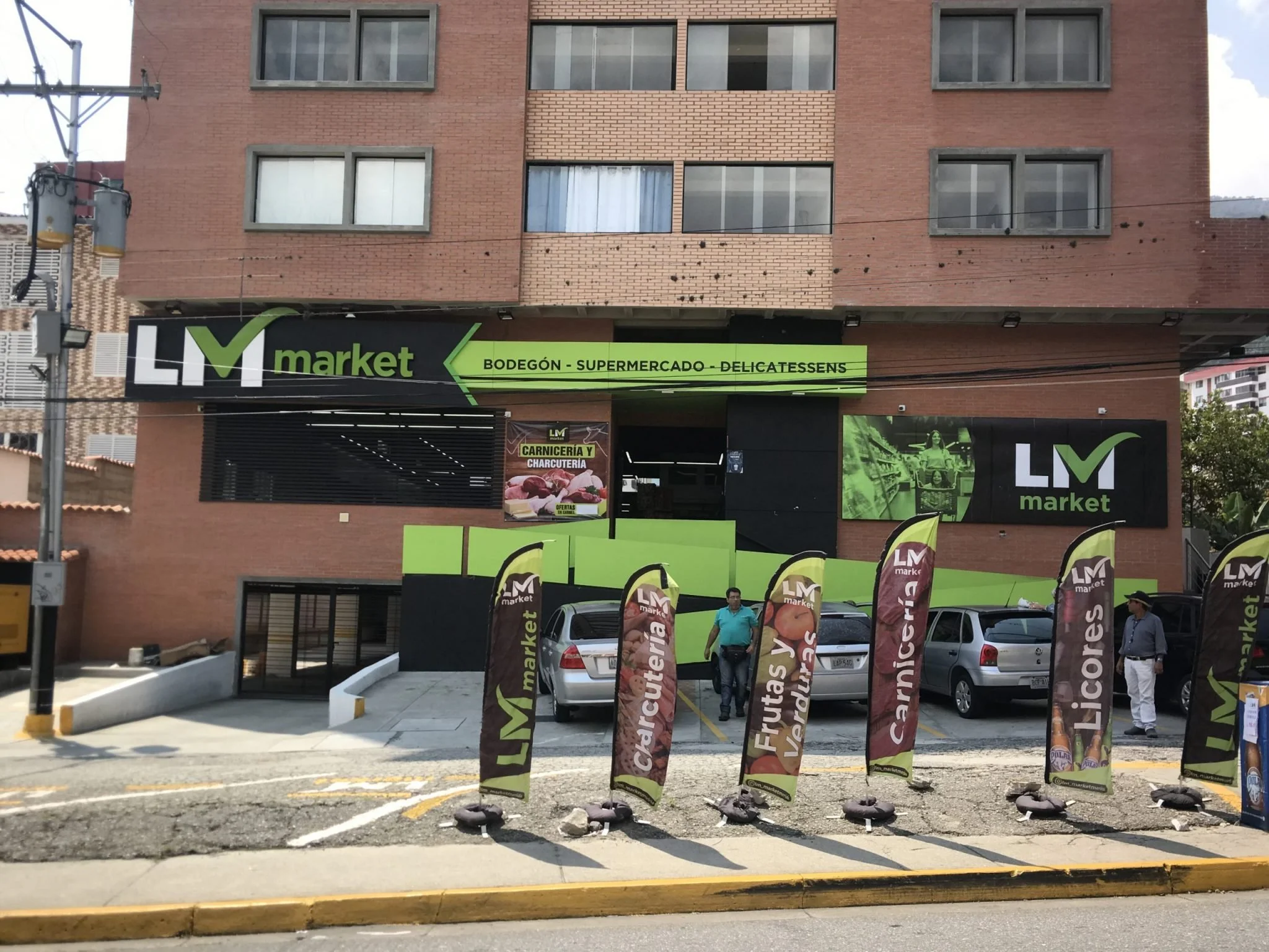 Exterior of a grocery store named LM Market with a brick building facade. Tall promotional flags are displayed in the parking lot, with trees and a street visible.