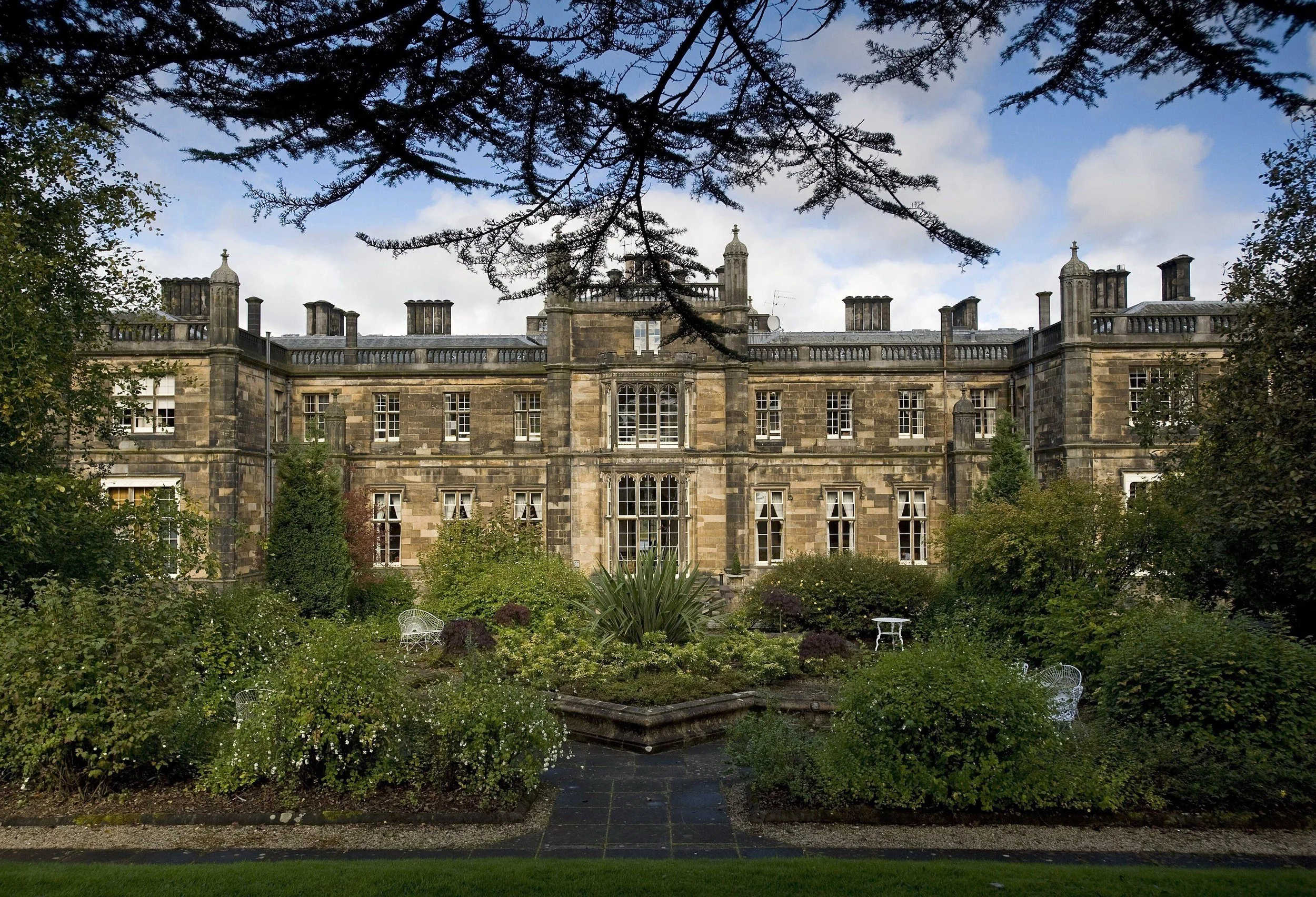 A large historic stone building with multiple windows, surrounded by lush greenery and trees in front, under a partly cloudy sky.