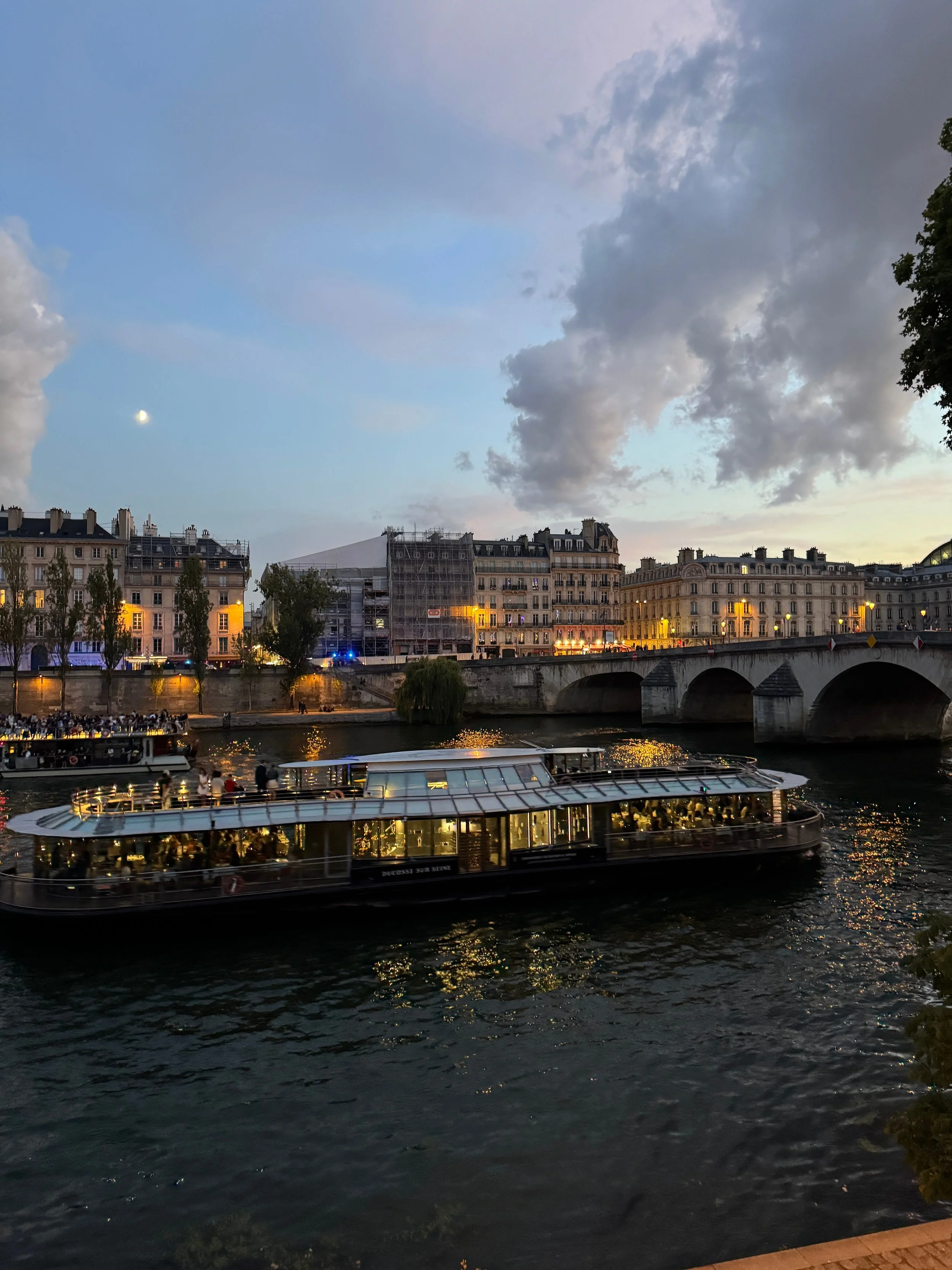 A river scene at dusk in Paris with a sightseeing boat on the water, historic buildings along the riverbank, and a partly cloudy sky with the moon visible.