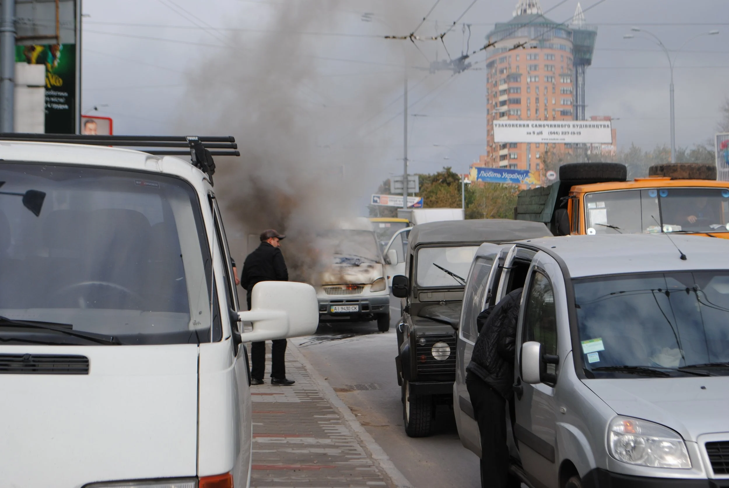 City street scene with multiple parked cars, smoking vehicle, and a man standing beside it. Tall buildings and overhead tram wires visible in the background. Kiev November 2011, Marina Parrish ©