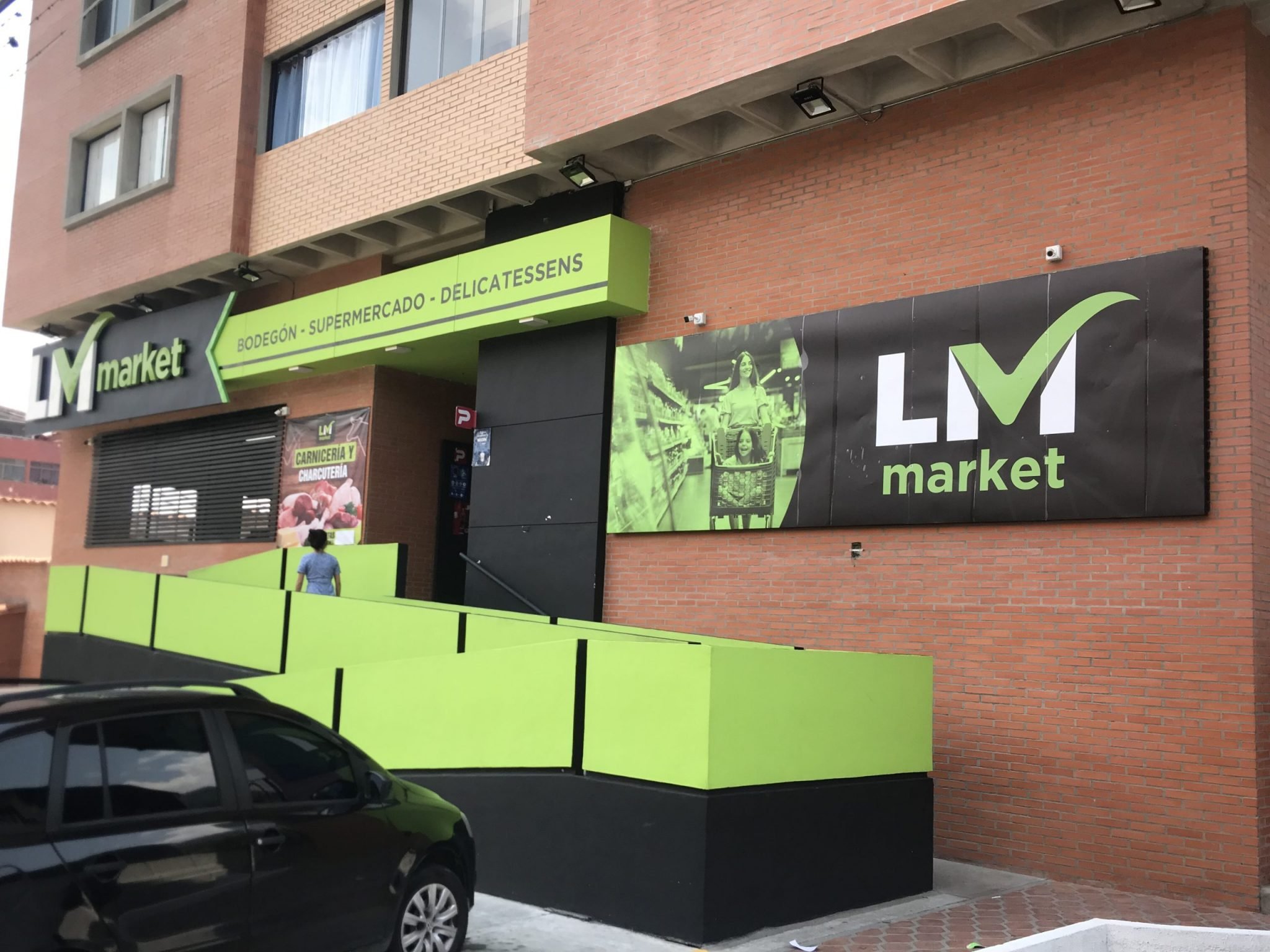 The exterior of a storefront with a green and black sign that reads 'LM market' and additional signage indicating it is a butcher shop, supermarket, and delicatessen. The building has a brick facade with windows. There is a person walking past, and c