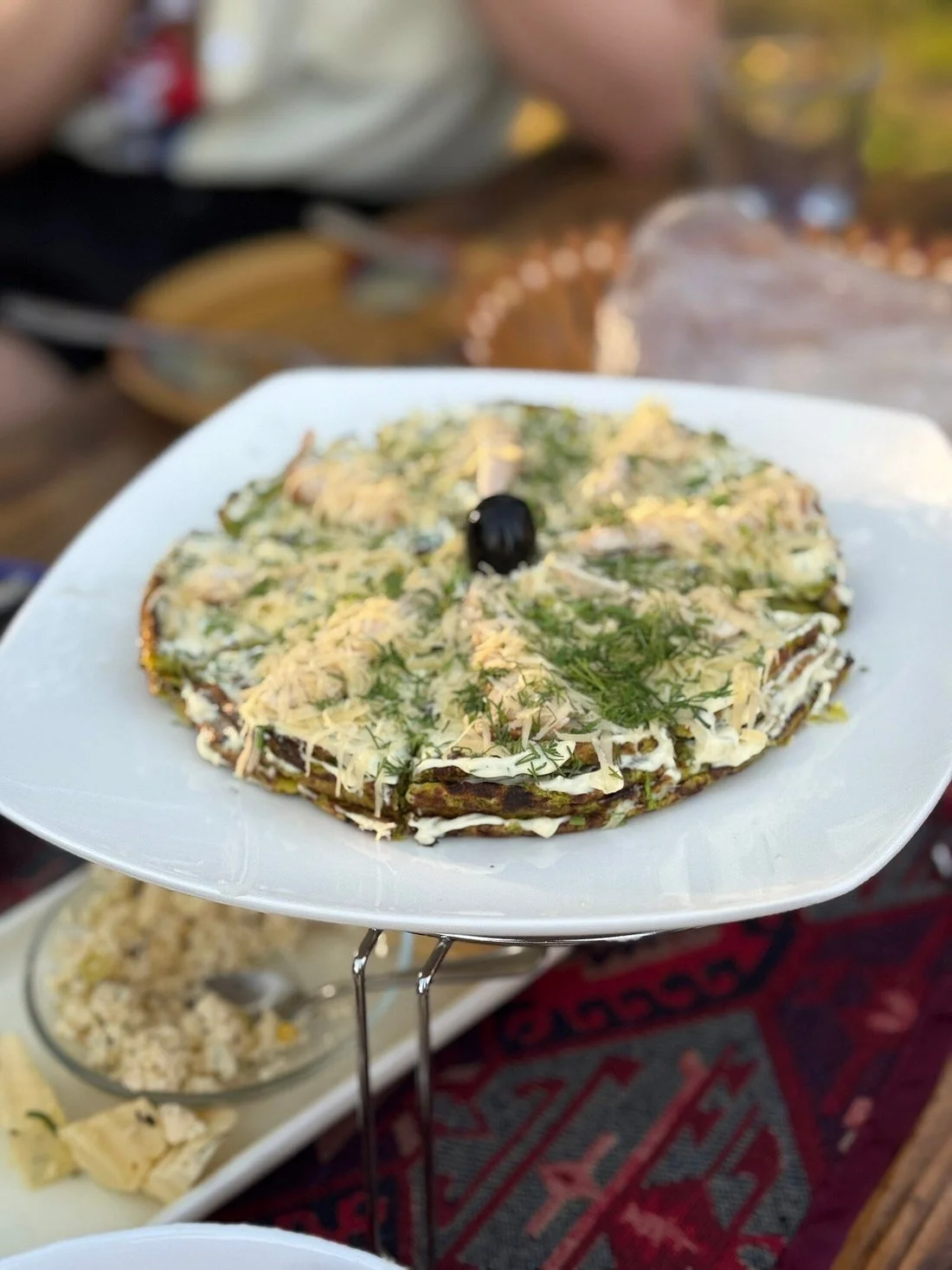 A plate of layered savory dish topped with cheese, herbs, and a black olive in the center, garnished with dill, served on a white square plate.