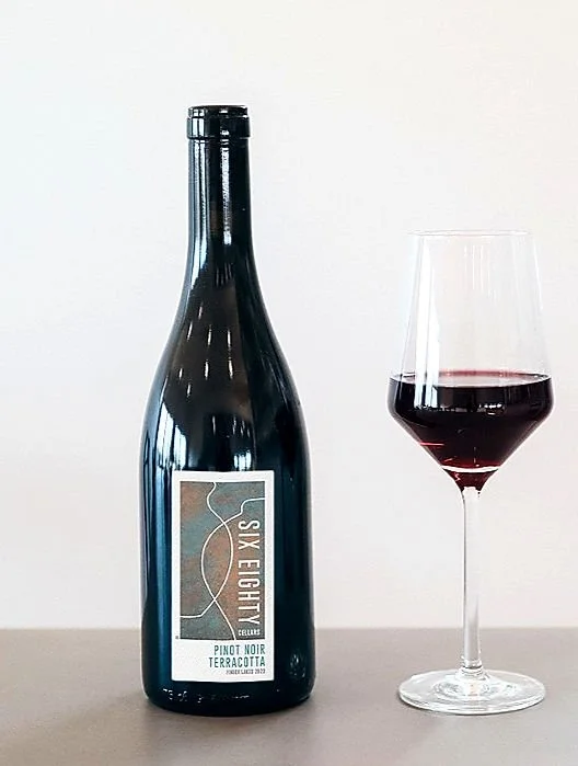 A bottle of Six Eighty Locals Pinot Noir Terrazzo wine next to a glass of red wine against a white background.