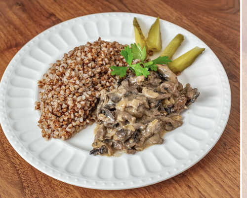 A white plate containing cooked buckwheat, a creamy mushroom stew garnished with a sprig of parsley, and slices of pickles, served on a wooden table.