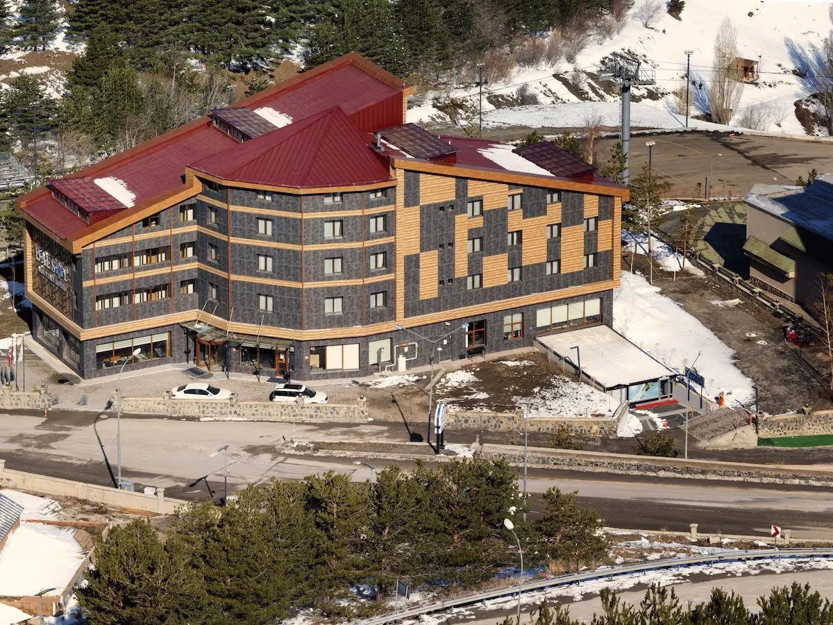 An aerial view of a modern multi-story building with a red roof, situated near a snow-covered landscape, trees, and a parking lot.