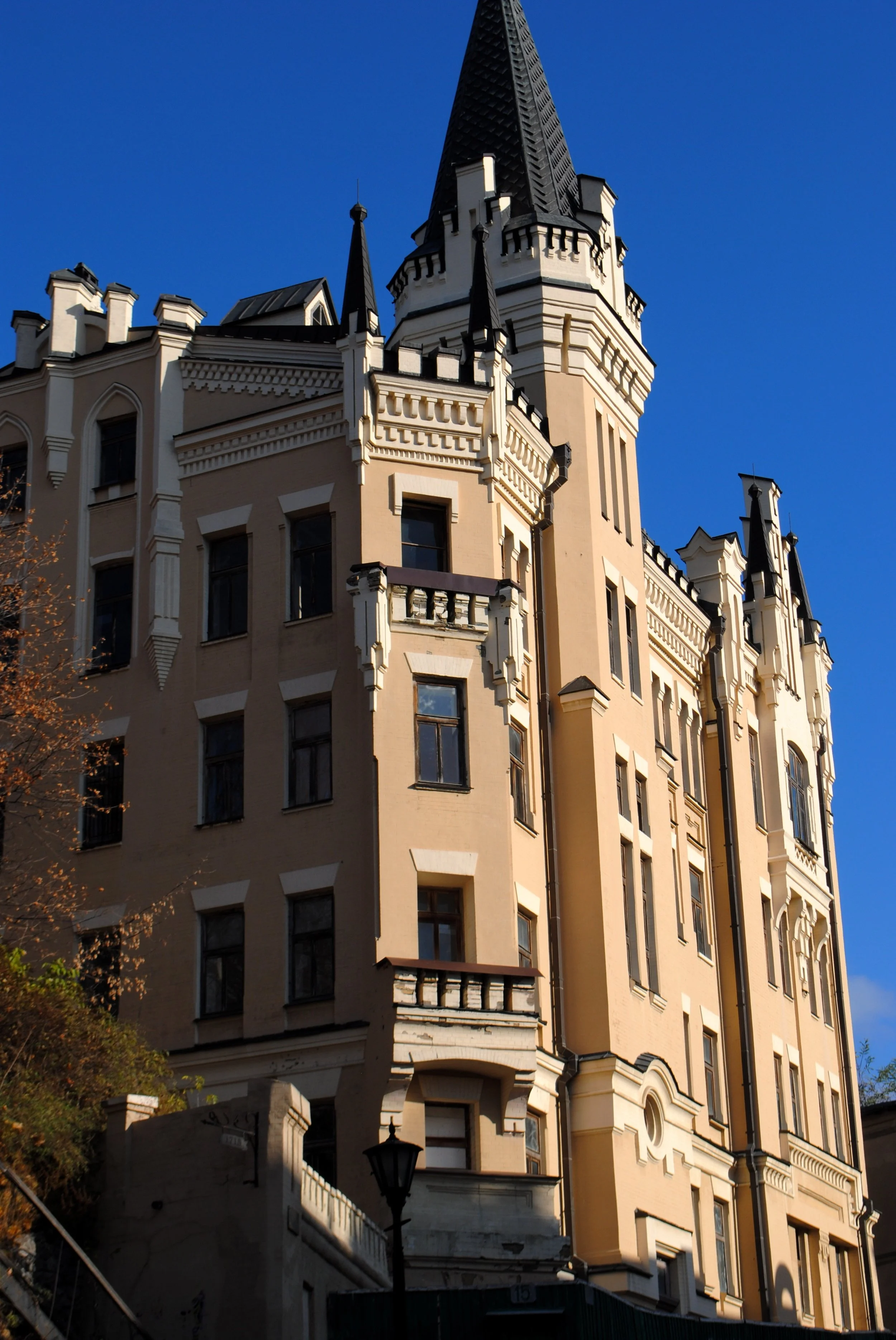 A tall, ornate building with a pointed spire and castle-like architecture, featuring multiple windows and decorative elements, set against a clear blue sky, with sunlight casting shadows on its cream-colored facade.