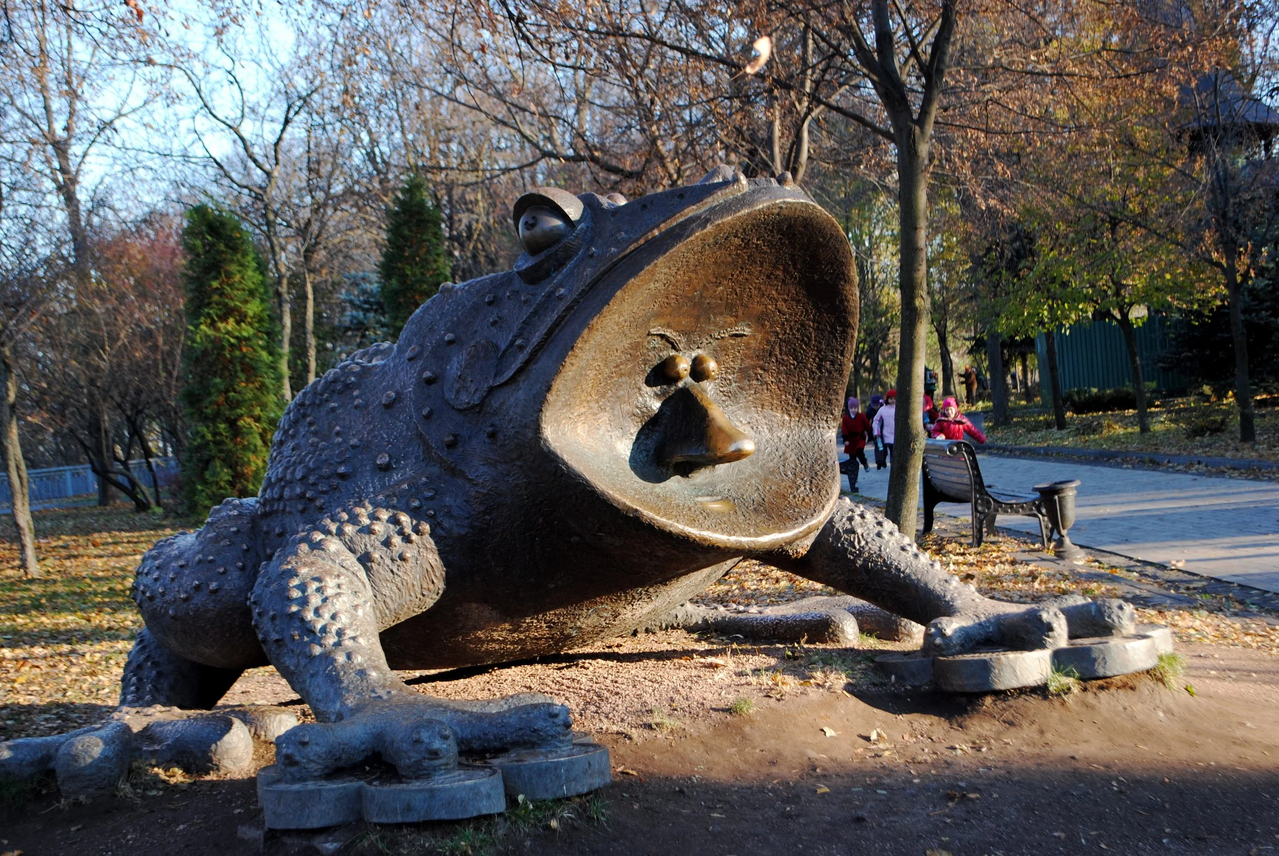 Bronze sculpture of a frog with exaggerated features, lying on the ground in a park with trees and children in the background.