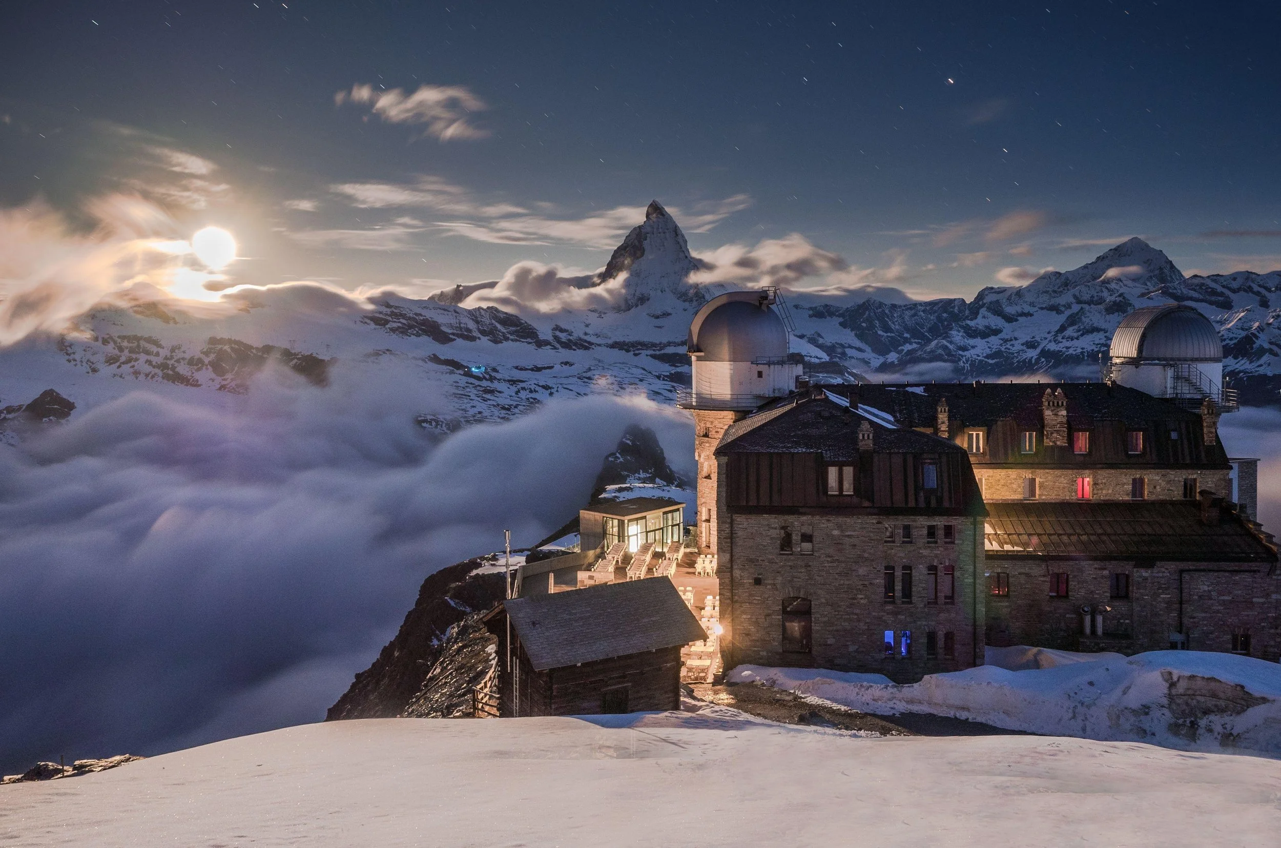 Nighttime view of a mountain observatory with domed telescopes, snow-covered peaks, and the Matterhorn in the background, illuminated by a bright moon.