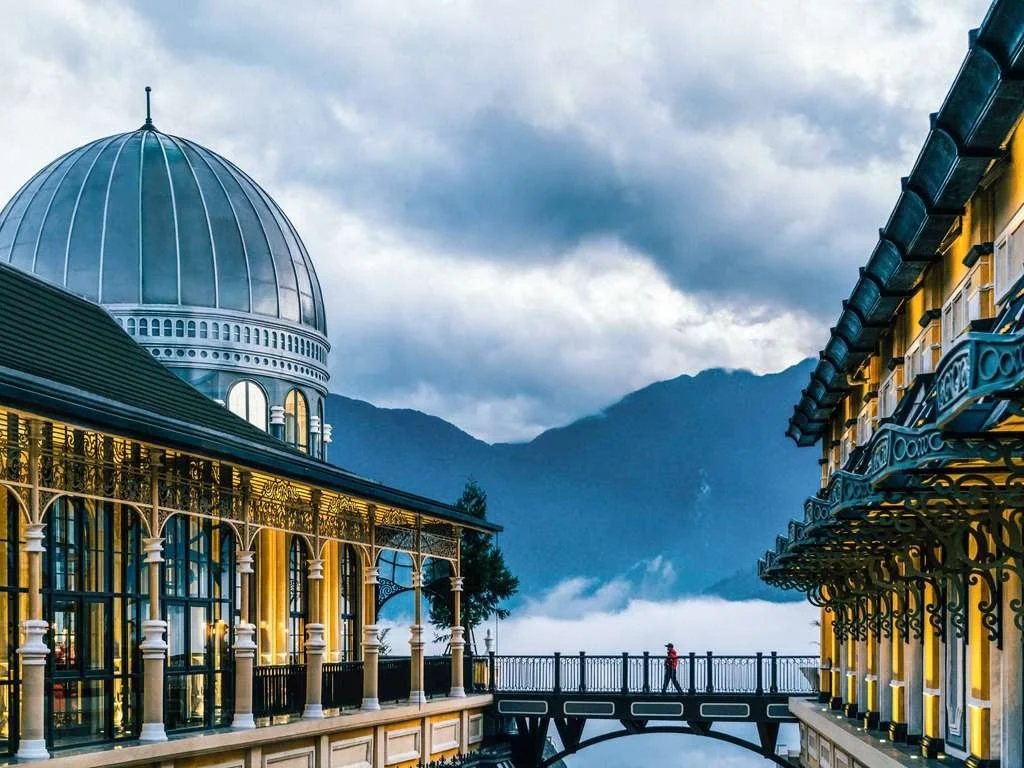Scenic view of a building with a large domed roof, ornate ironwork on the balconies, and a person walking on a bridge against a backdrop of mountains and cloudy sky.
