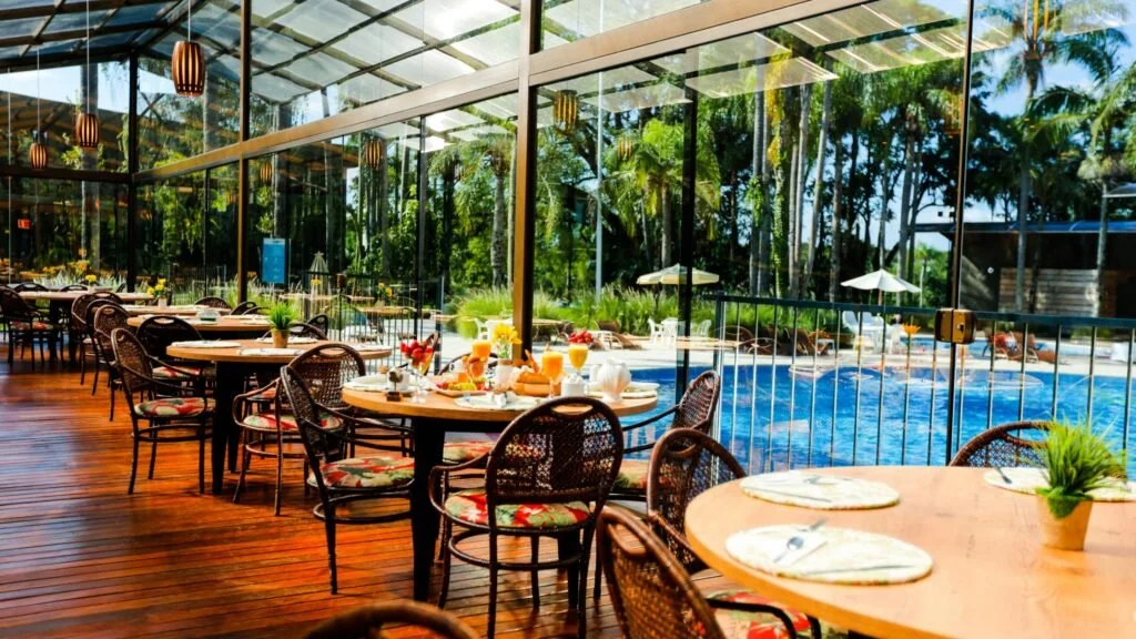 Indoor dining area with round tables set for breakfast, glass walls, and outdoor pool view with sun umbrellas and lounge chairs.