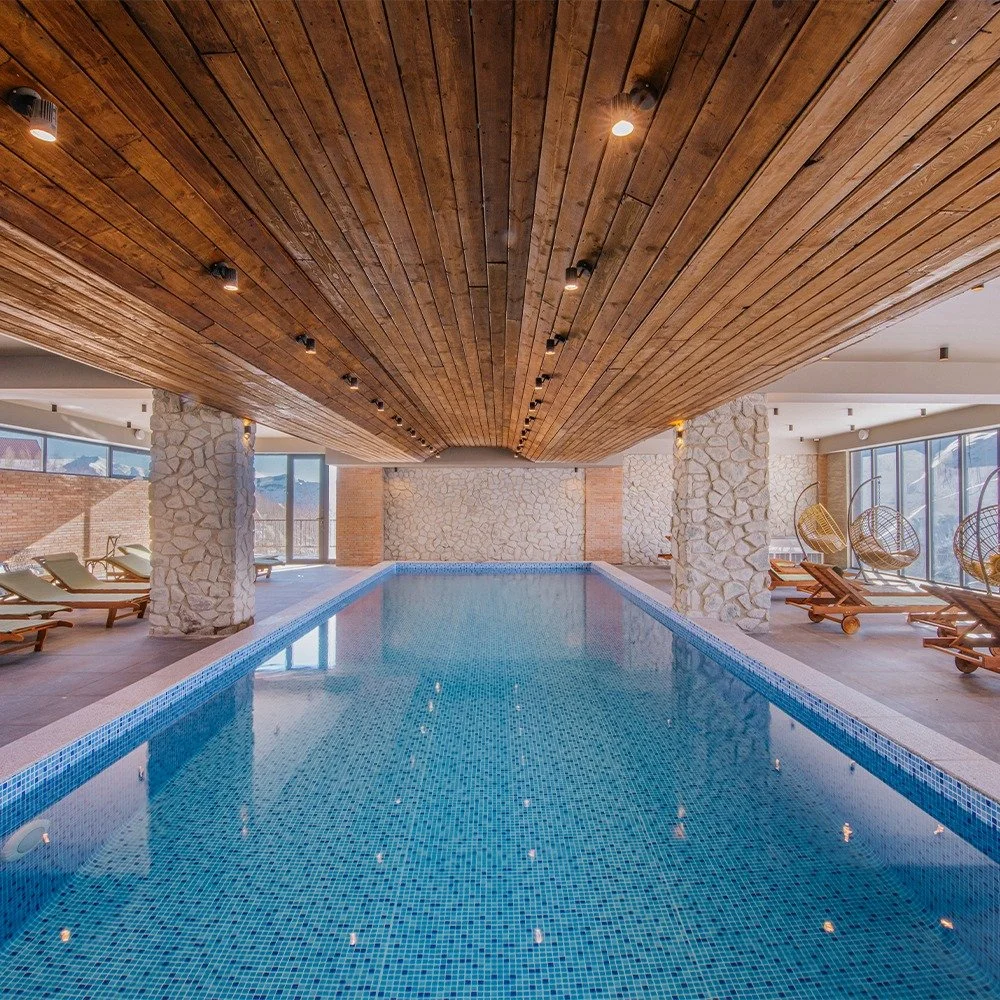 Indoor swimming pool area with blue tiled pool, stone pillars, wooden ceiling with recessed lighting, large windows, and lounge chairs.