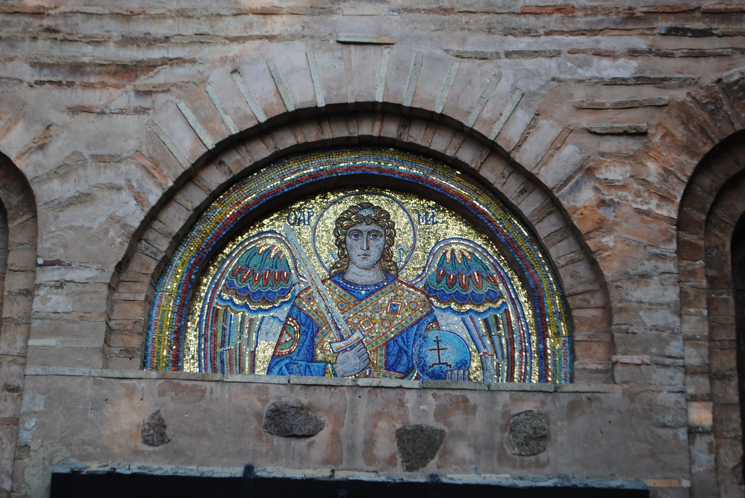 Colorful mosaic of a religious figure, likely an angel or saint, with a halo, set in a stone wall arch.