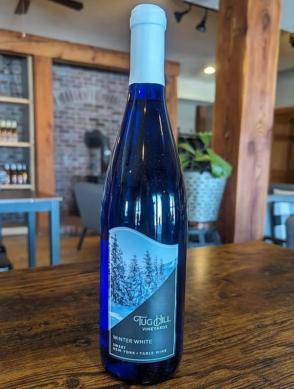 A blue glass wine bottle with a winter landscape label sitting on a wooden table in a cozy indoor setting.