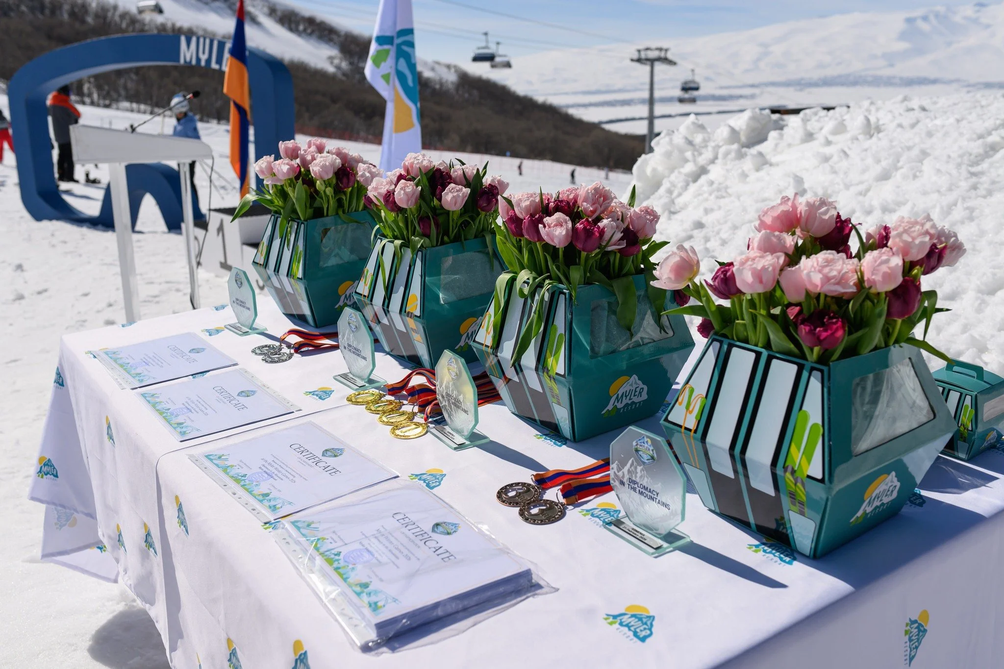 Awards table with medals, certificates, and pink, purple, and white tulip bouquets, set on snow at the Myler Ski Area during a sunny winter day with ski lifts in the background.