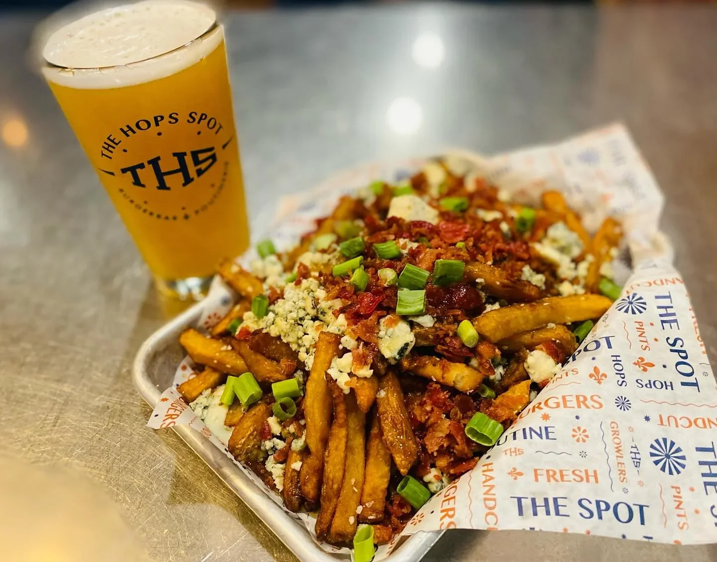 Basket of loaded fries topped with cheese, green onions, bacon, and crumbled cheese, with a pint of beer in a glass labeled 'THE HOPS SPOT' in the background.
