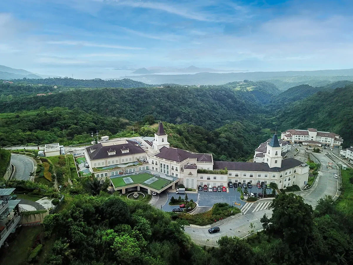 A large, white castle-like building with towers and a parking lot, surrounded by lush green hills and mountains under a partly cloudy sky.