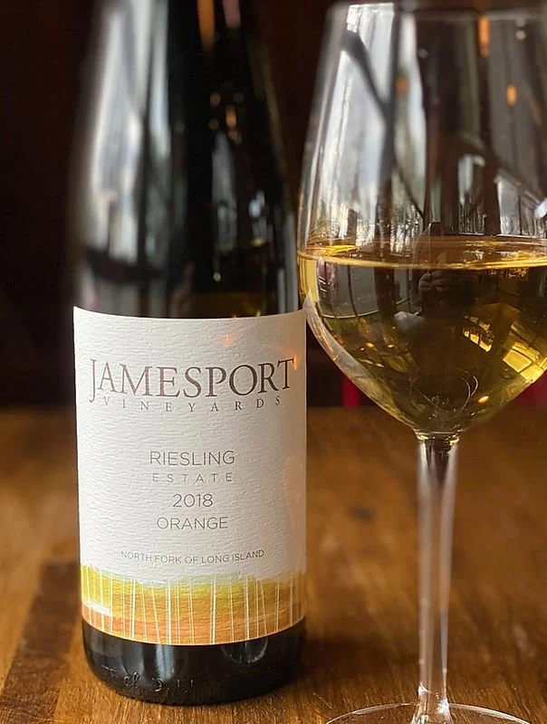 A bottle of Jamesport Vineyards Riesling 2018 Orange wine next to a glass filled with white wine on a wooden table.