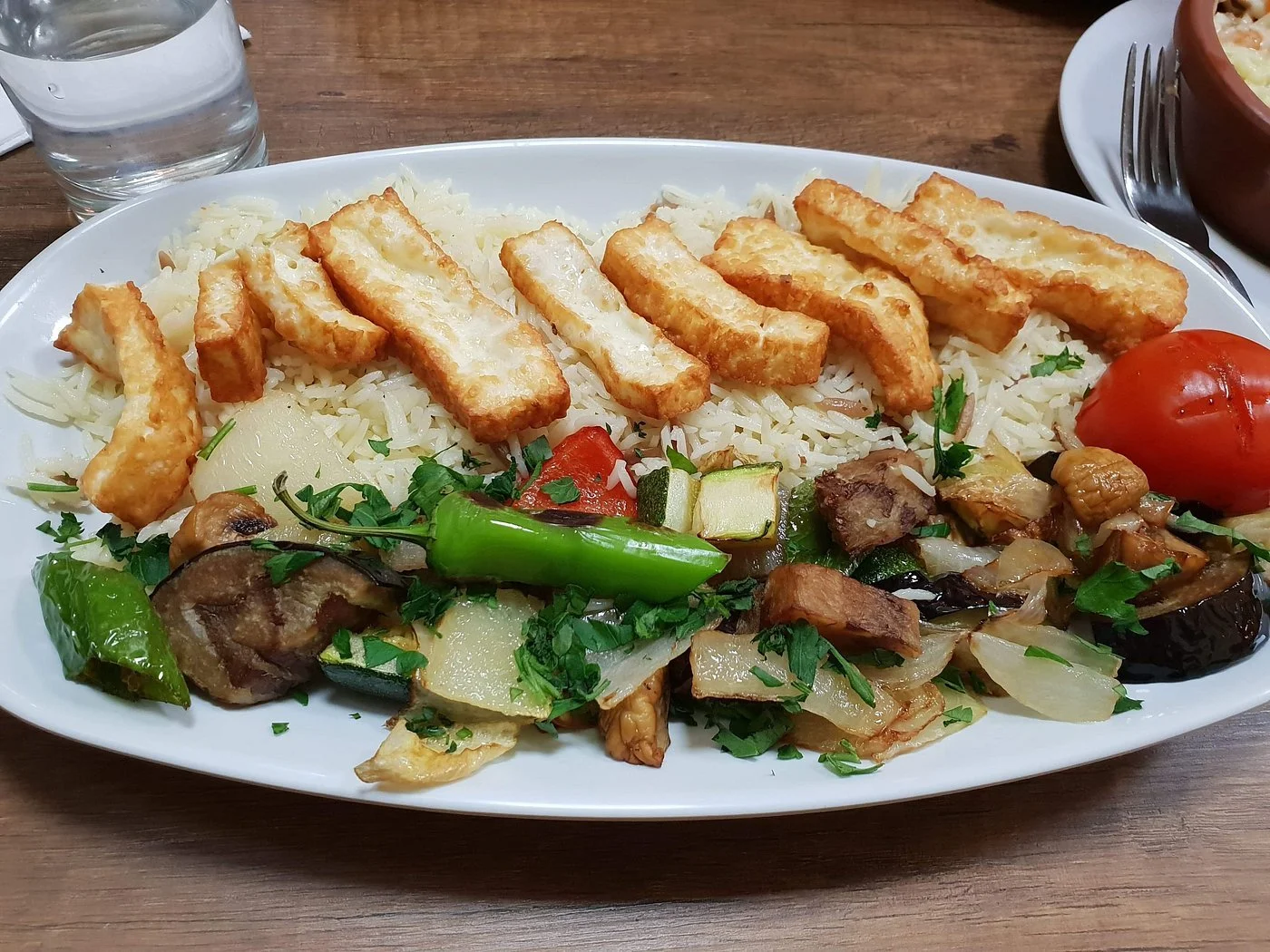Plate of rice with fried chicken strips, vegetables including zucchini, eggplant, onion, and a whole roasted tomato, garnished with chopped herbs.