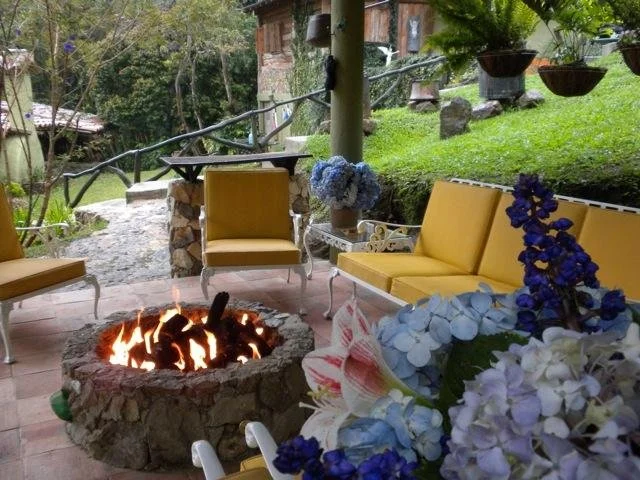 Outdoor patio area with yellow cushioned seating, a stone fire pit with a fire, and hanging flower pots with purple and white flowers, surrounded by greenery and a wooden house in the background.