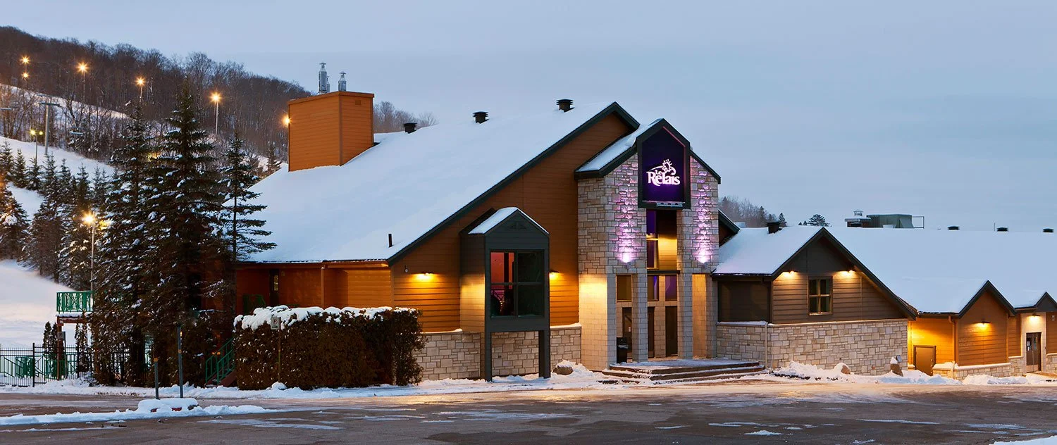 A snow-covered building with a lit sign that reads 'Rebals,' surrounded by snow and trees, with a hill and streetlights in the background, during twilight or evening.