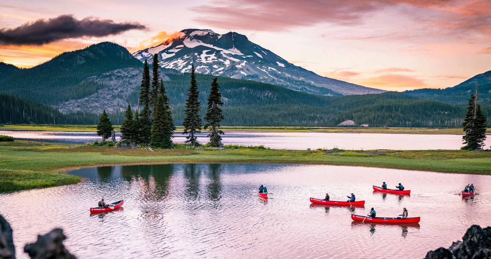 Six people kayaking in a calm lake surrounded by green trees and mountains with a snow-capped volcano in the background during sunset.