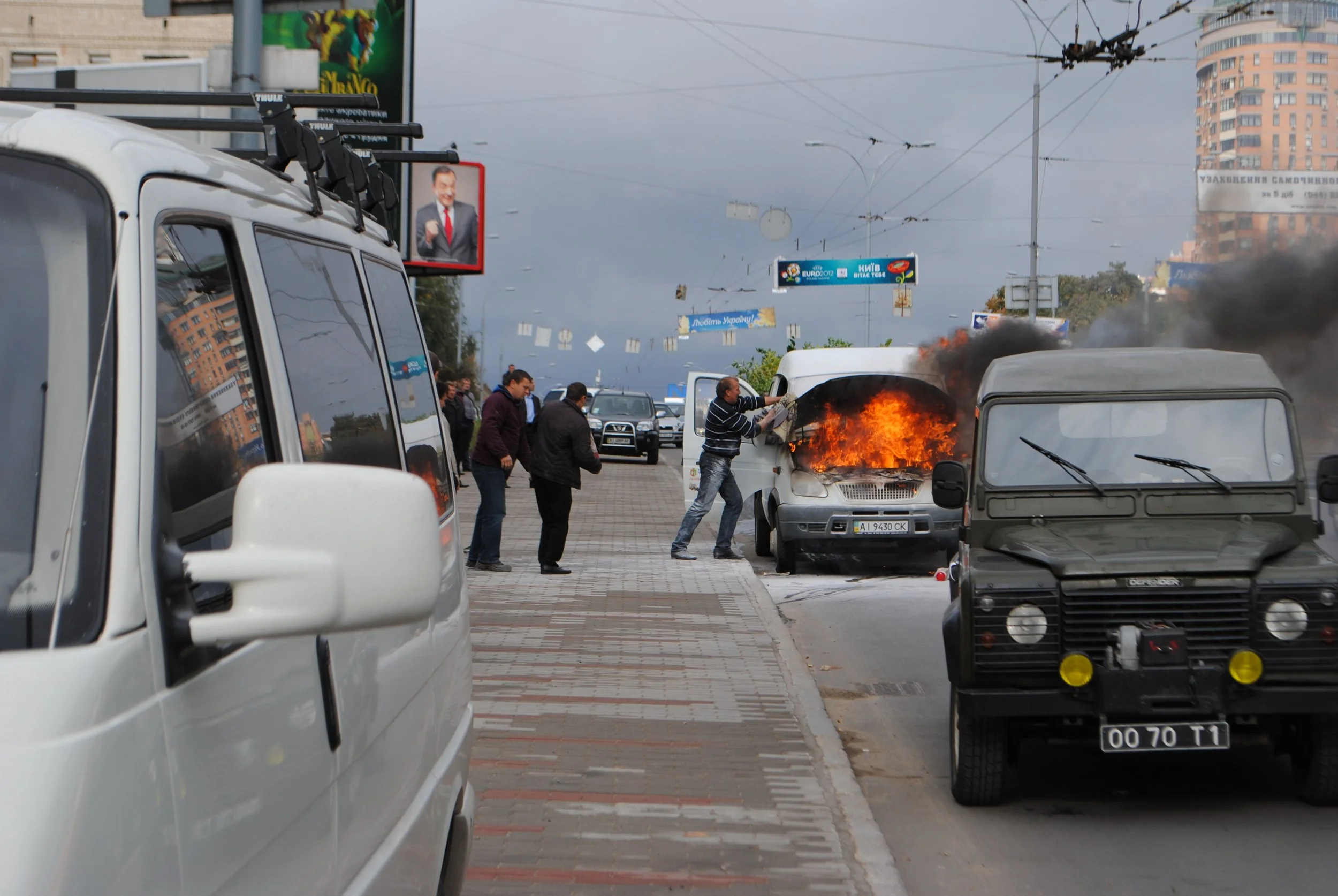 A car is on fire in a busy city street while people gather nearby, and a man is seen throwing an object into the flames. Multiple vehicles are parked and moving around, with buildings and billboards in the background.