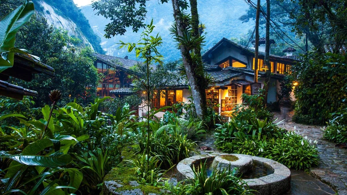 A lush, tropical garden path leading to a cozy house with warm interior lights, surrounded by dense greenery and tall trees in a mountainous area.