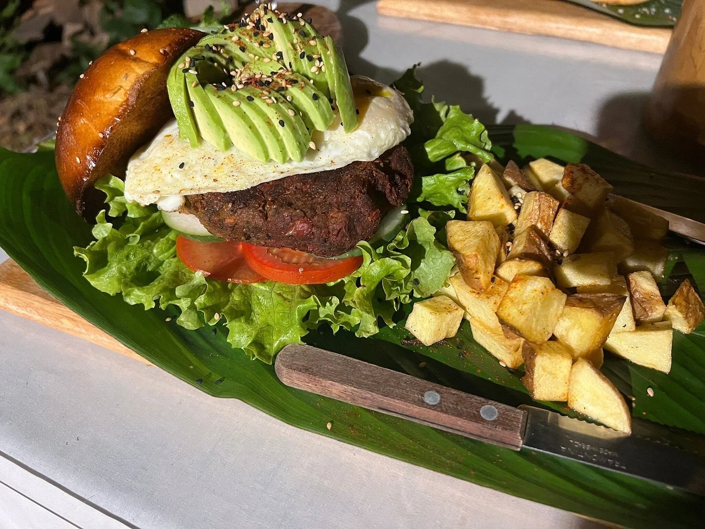 A burger with lettuce, tomato, a beef patty, melted cheese, topped with sliced avocado, a fried egg, and a toasted bun, served with roasted cubed potatoes on a banana leaf-lined tray.