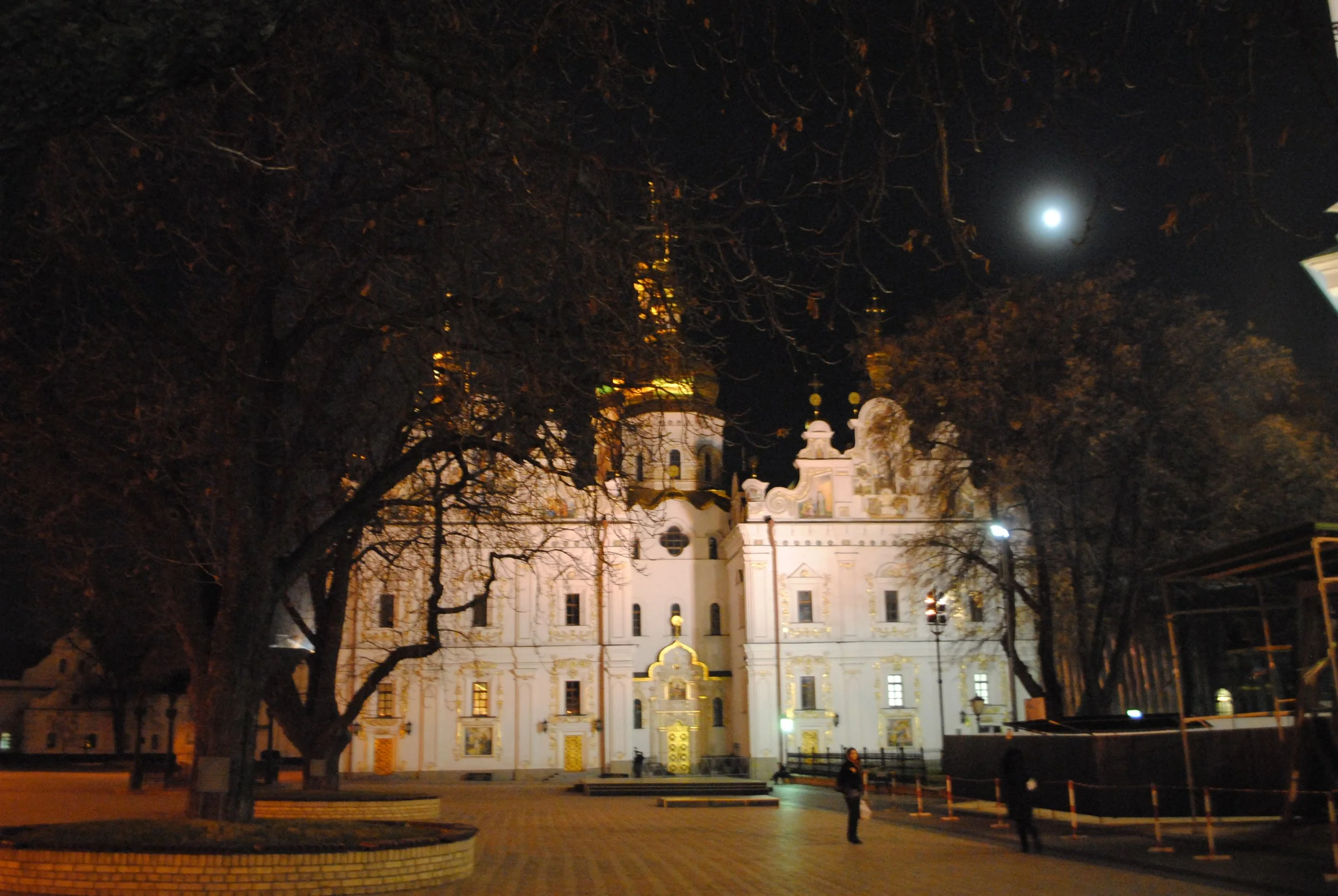 Nighttime scene of a historic white building with ornate architectural details, illuminated by exterior lights, framed by leafless trees and a visible full moon in the dark sky.