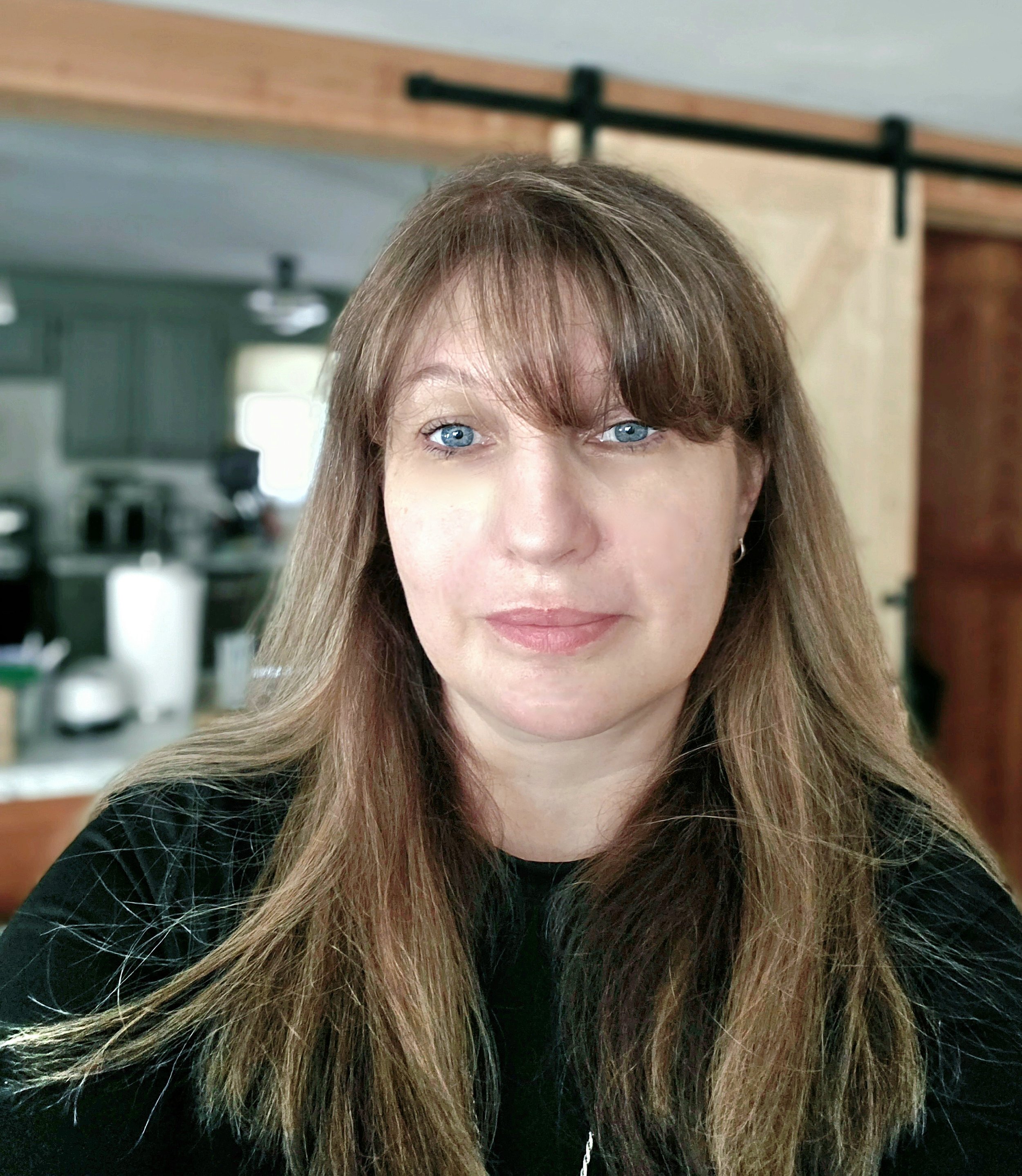 Close-up of a woman with long brown hair and blue eyes, wearing a black top, in a home setting with kitchen visible in background.