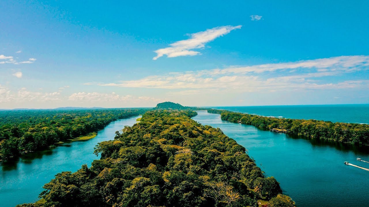 Aerial view of a narrow strip of land covered in dense green trees, dividing two large bodies of water, with a distant island on the horizon and a blue sky with scattered clouds.