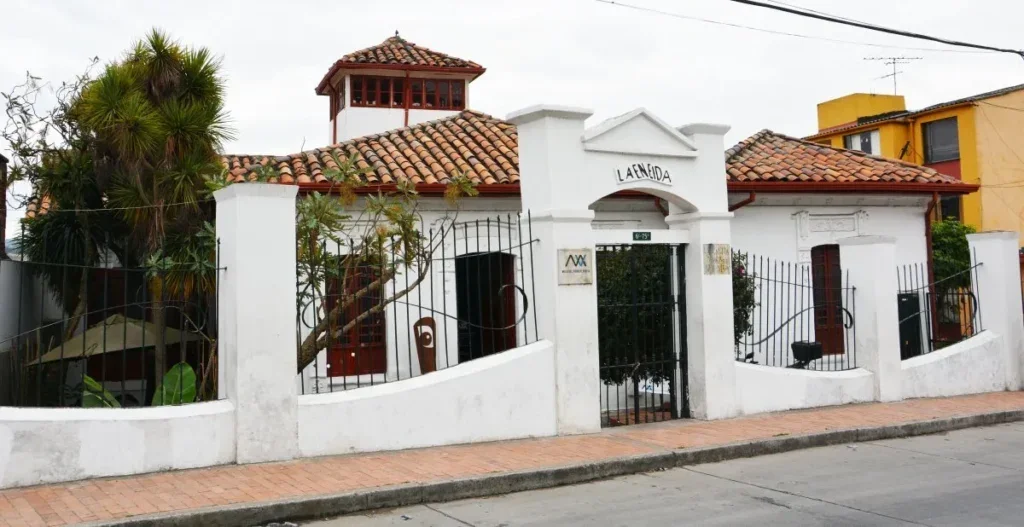 A white house with a red tiled roof, a small tower, and a gated entrance, surrounded by a white curved wall and iron fence, with trees and yellow building in the background.