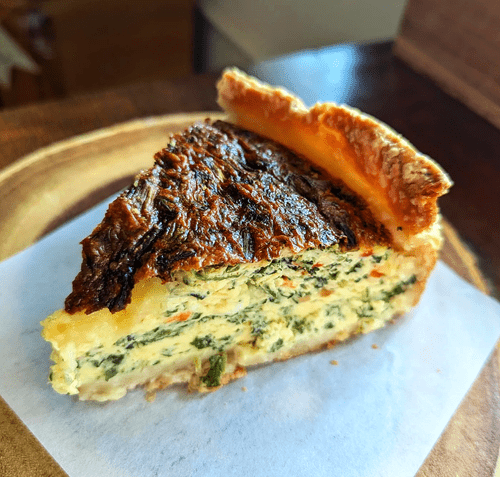 A slice of spinach and cheese quiche with a flaky crust, served on white paper on a wooden surface.