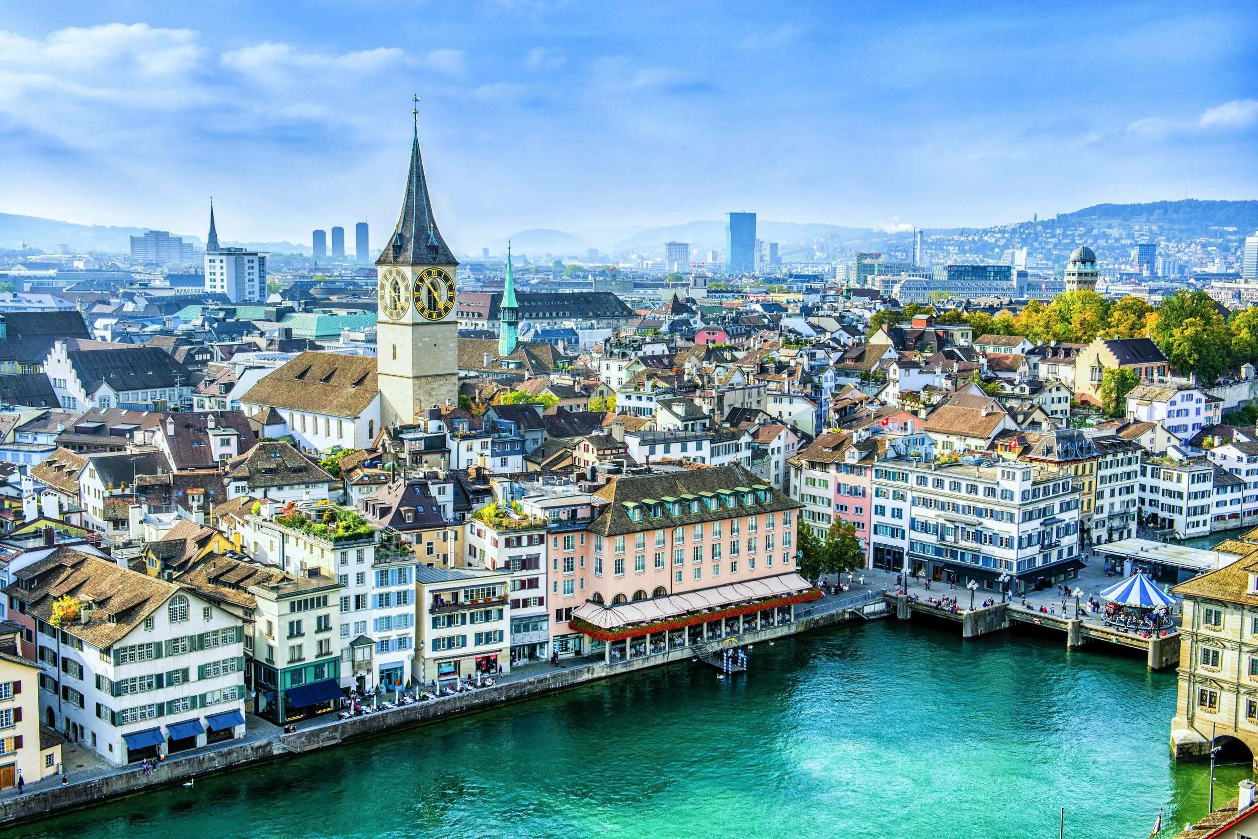 Green valley with a stream, trees, small buildings, and mountains in the background under cloudy sky