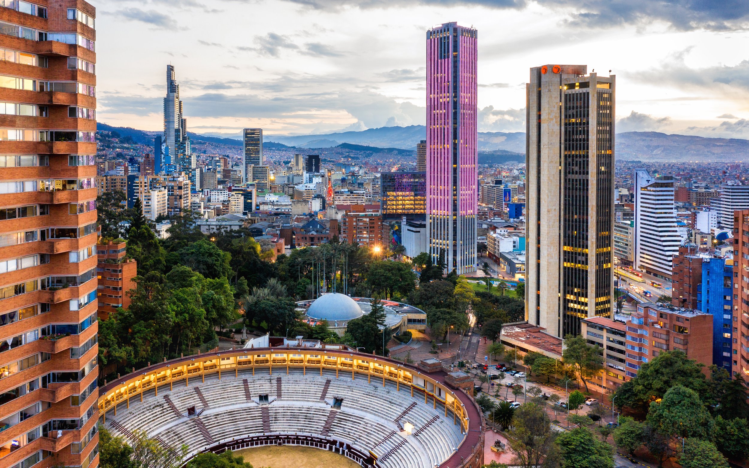 City skyline with tall modern skyscrapers, mountains in the background, and an open-air theater with tiered seating in the foreground during dusk.