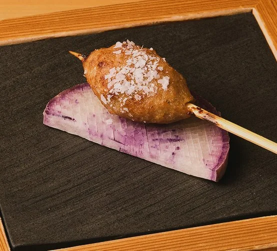 A Japanese skewered croquette on a stick resting on a slice of purple radish on a black tray with a wooden border.