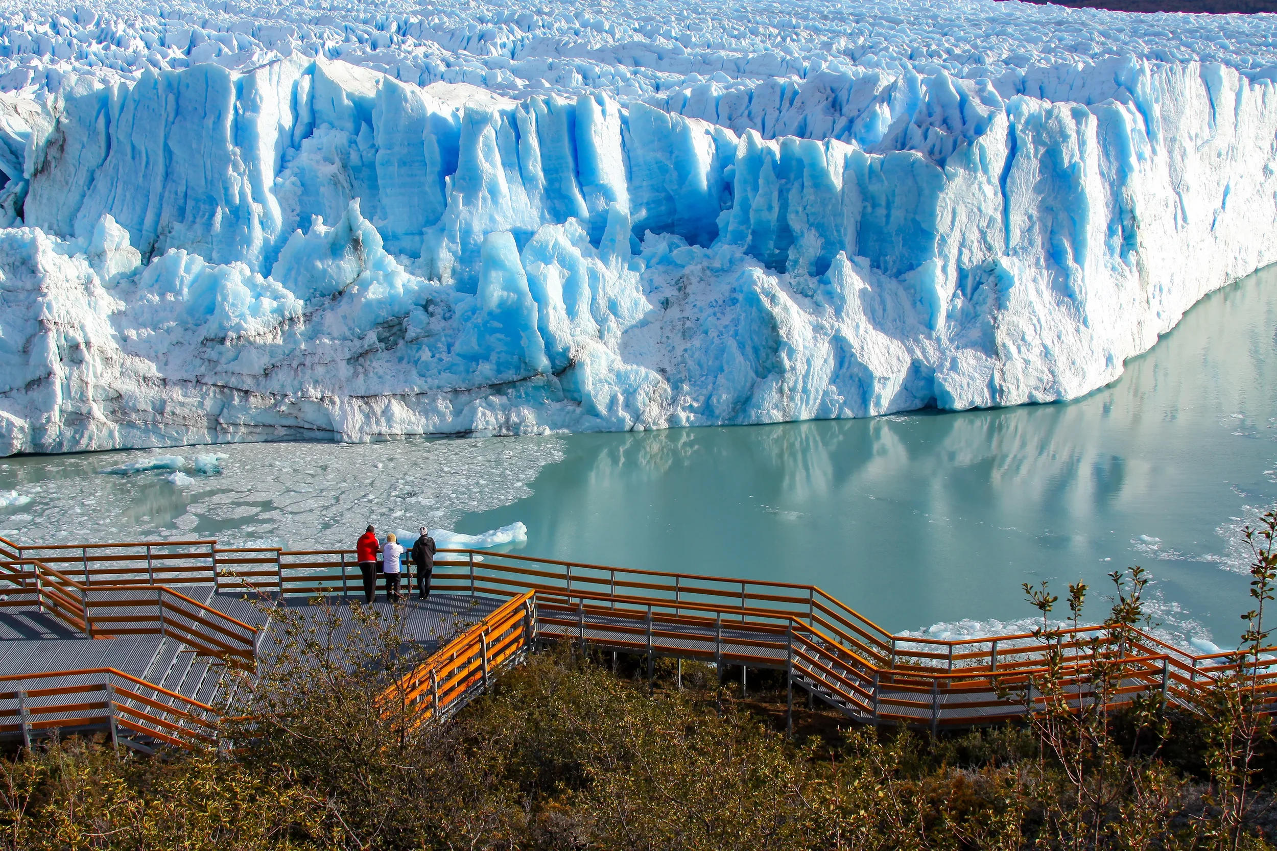 People viewing a large glacier calving into a body of water from an observation deck with orange railings.
